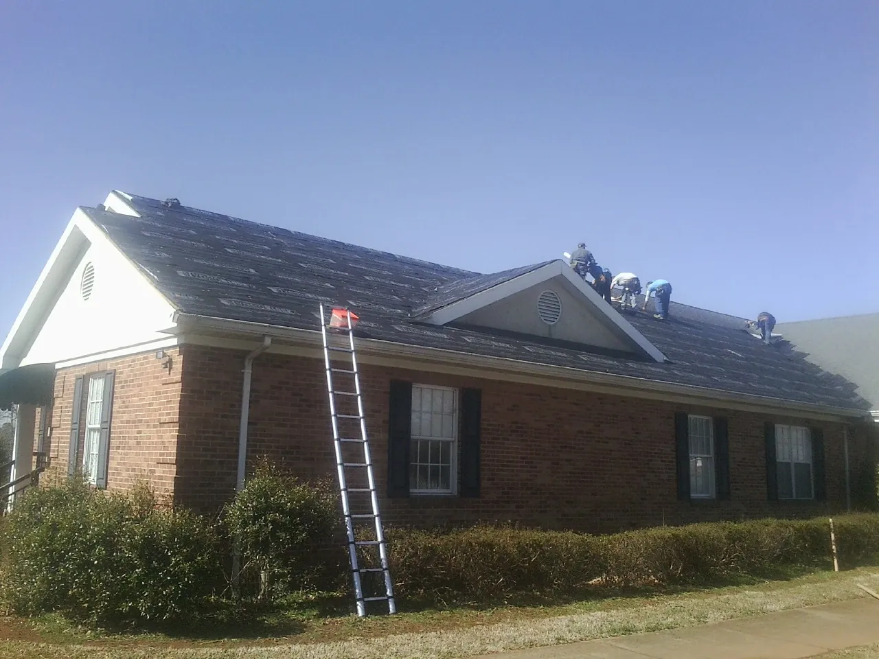 Crew tearing shingles off a brick ranch roof with a ladder set — Emergency Roof Repair work by our crew in Calvin