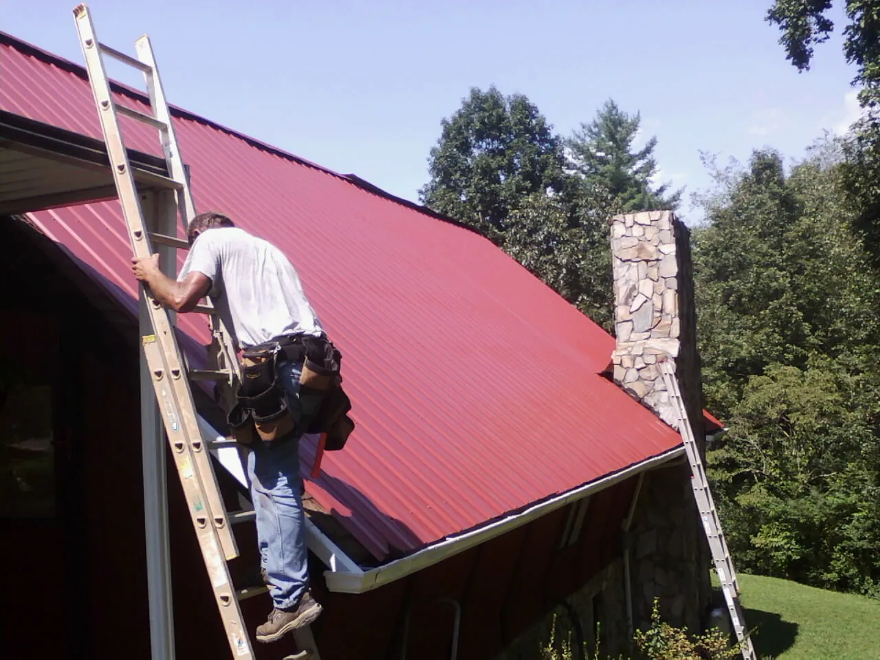 Roofer on a ladder beside a red metal roof and stone chimney — Soffit & Fascia Repair by Roofing Brothers in Calvin